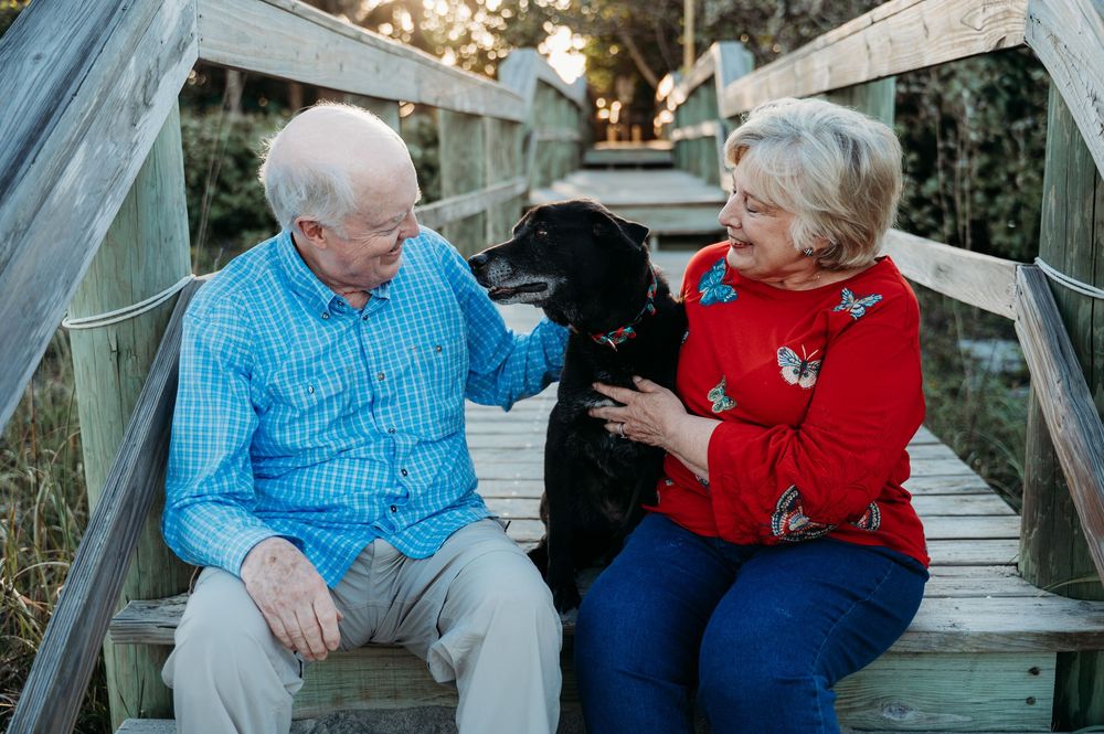 Elderly couple sitting on wooden bridge petting their black Labrador dog outdoors in a warm family portrait.