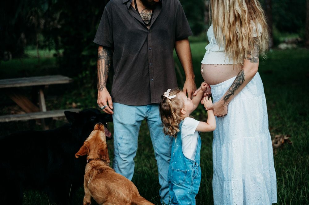 Tattooed couple with pregnant woman in white dress, young girl, and brown dog posing outdoors in green park setting.