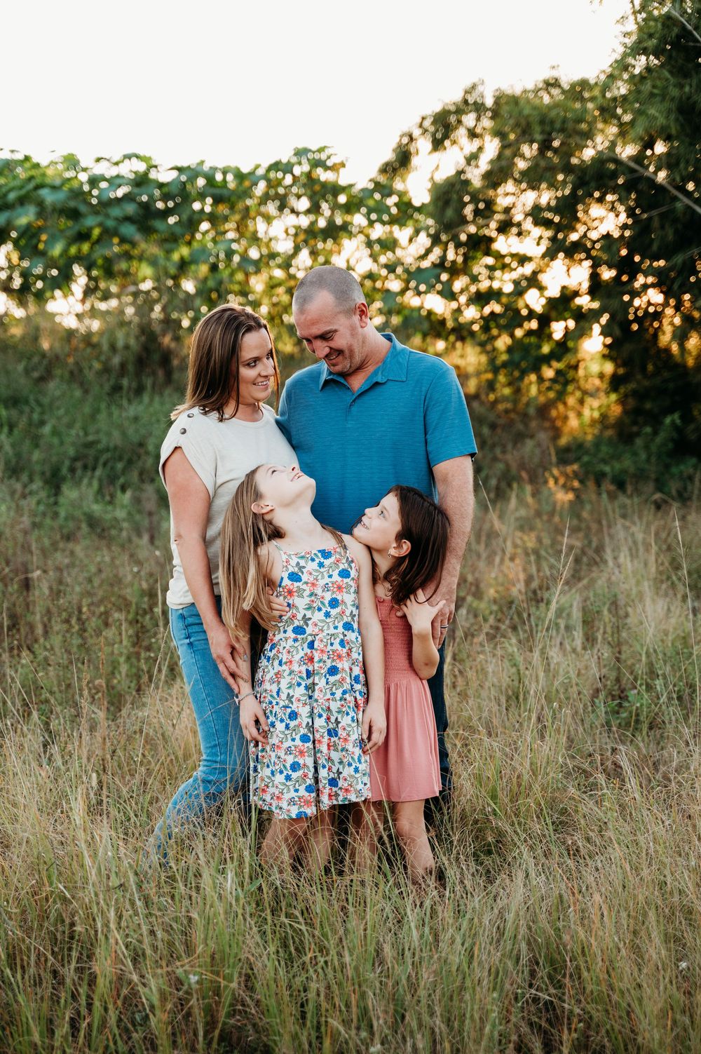 Family of four smiling together in a golden sunlit field, two girls looking up at their parents with joy.