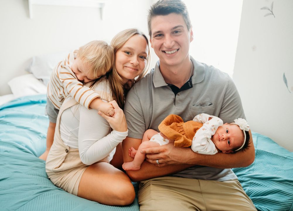 Young family poses on bed with toddler and newborn baby, smiling warmly in bright white room.