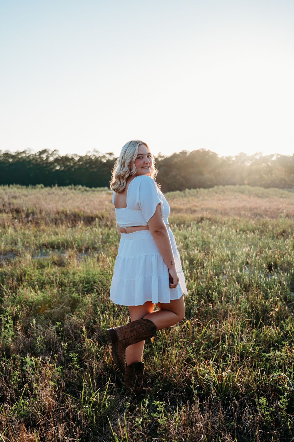 Young blonde woman in white crop top and skirt with brown cowboy boots standing in a sunlit green field.