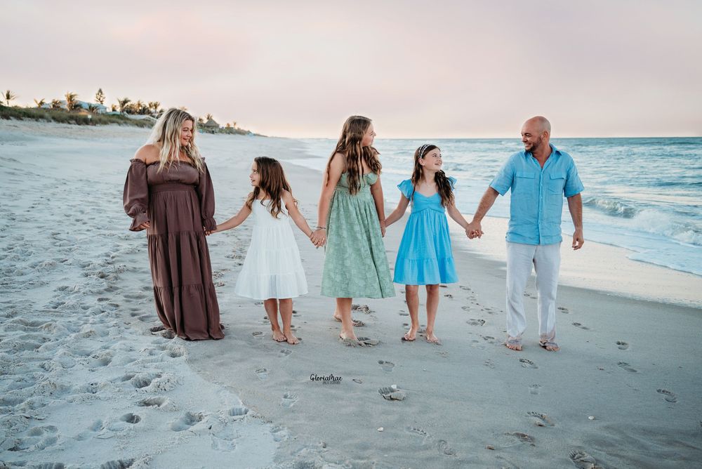 Family of five holding hands walking on a sandy beach at sunset, wearing colorful summer dresses and casual attire.