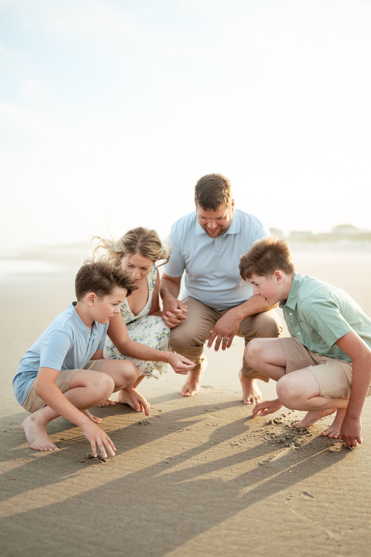 Family of four crouching together examining something in the sand on a sunny beach.