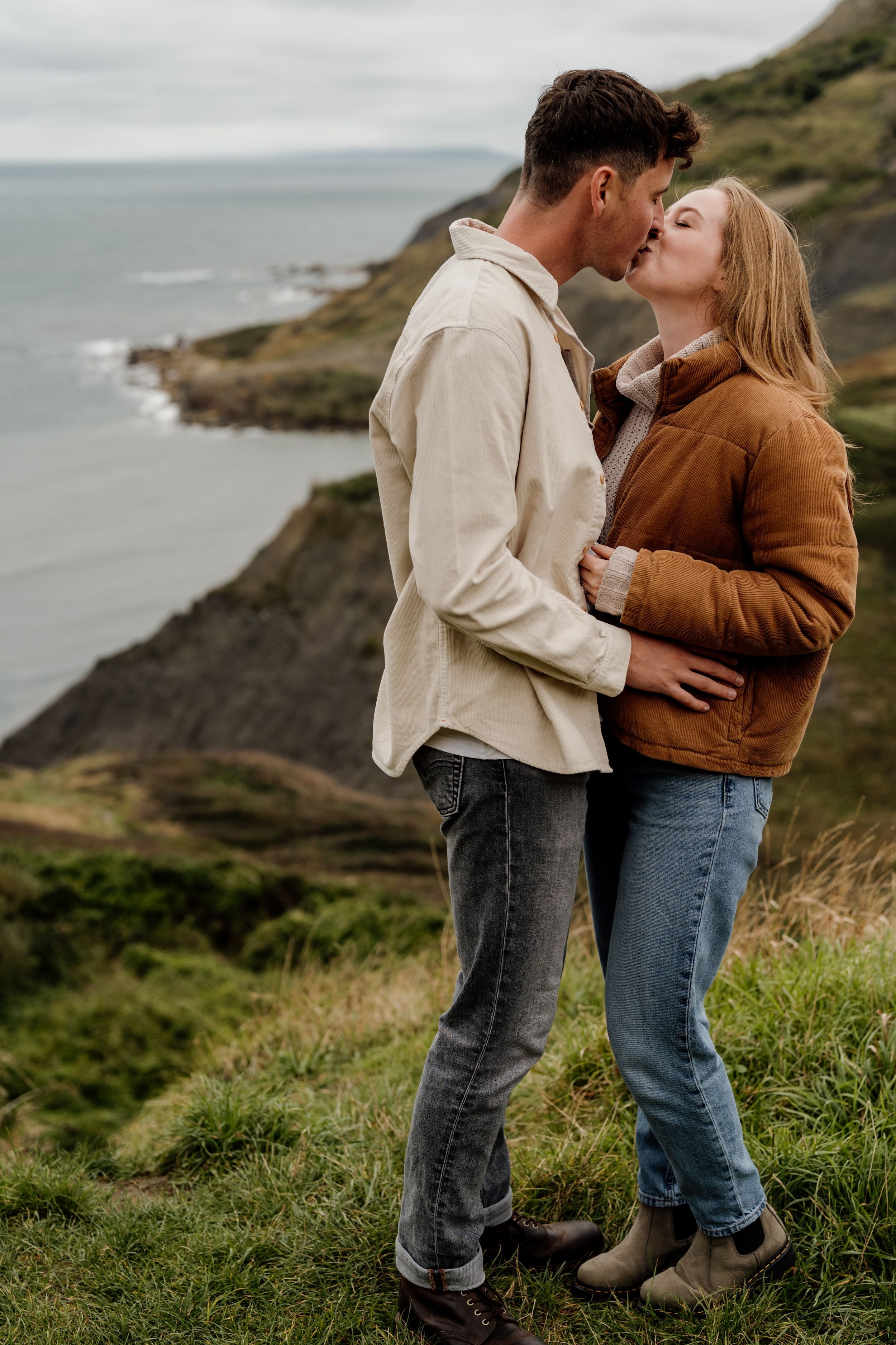 Couple sharing a romantic kiss on a coastal cliff, dressed in casual autumn outfits with ocean views behind them.