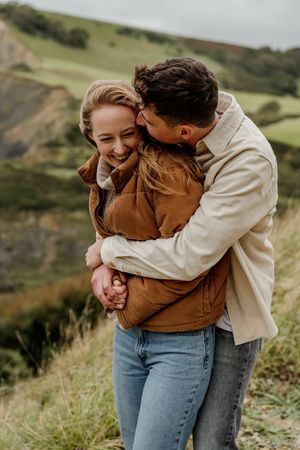 A couple embraces outdoors, woman laughing joyfully as man kisses her cheek, green hills in background.