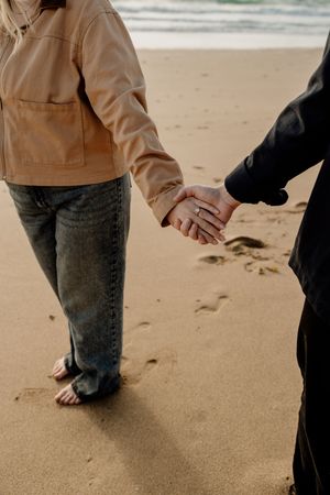 Couple holding hands on sandy beach, woman wearing engagement ring, tan jacket and jeans, walking barefoot near ocean waves.