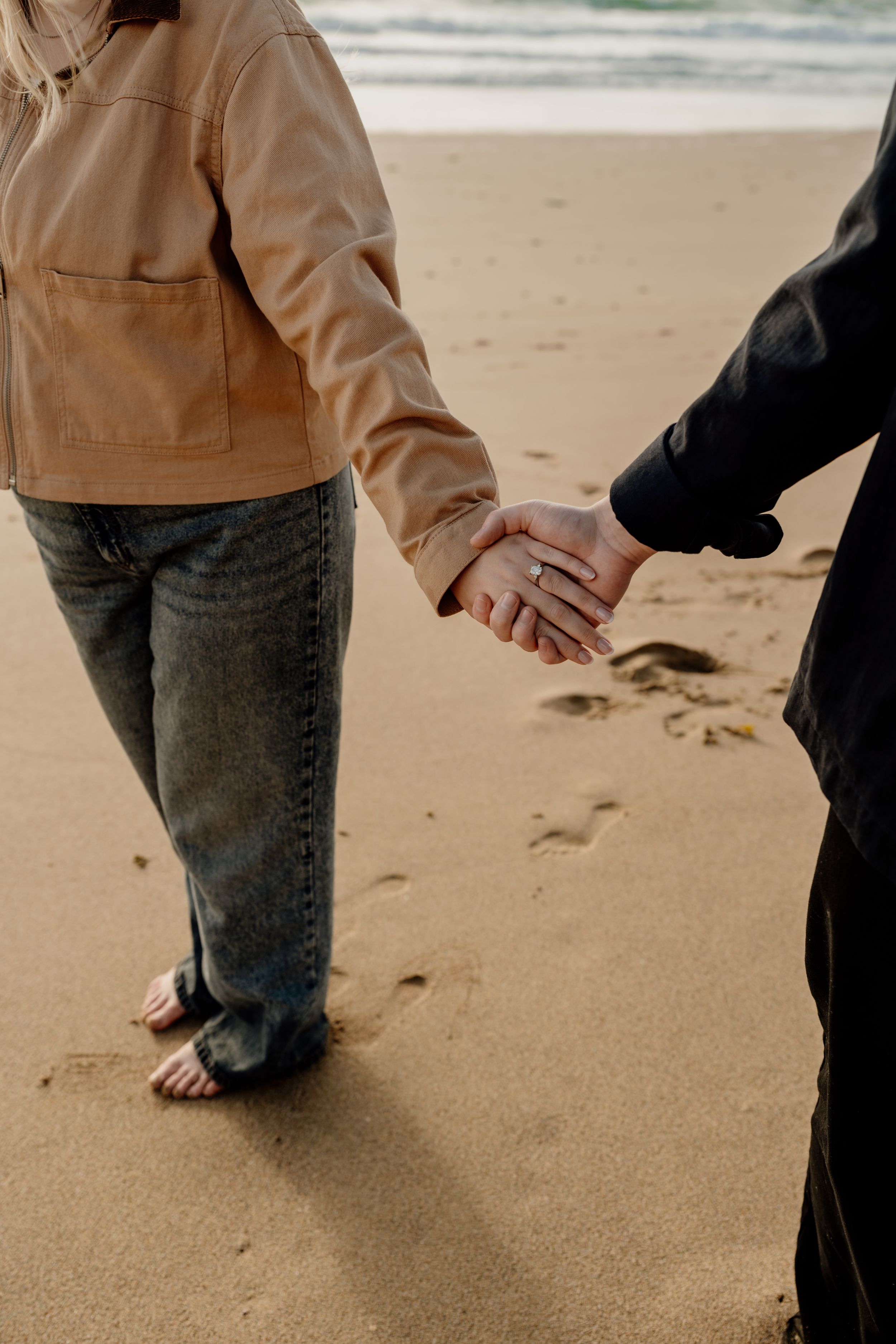 Couple holding hands on sandy beach, woman wearing engagement ring, tan jacket and jeans, walking barefoot near ocean waves.