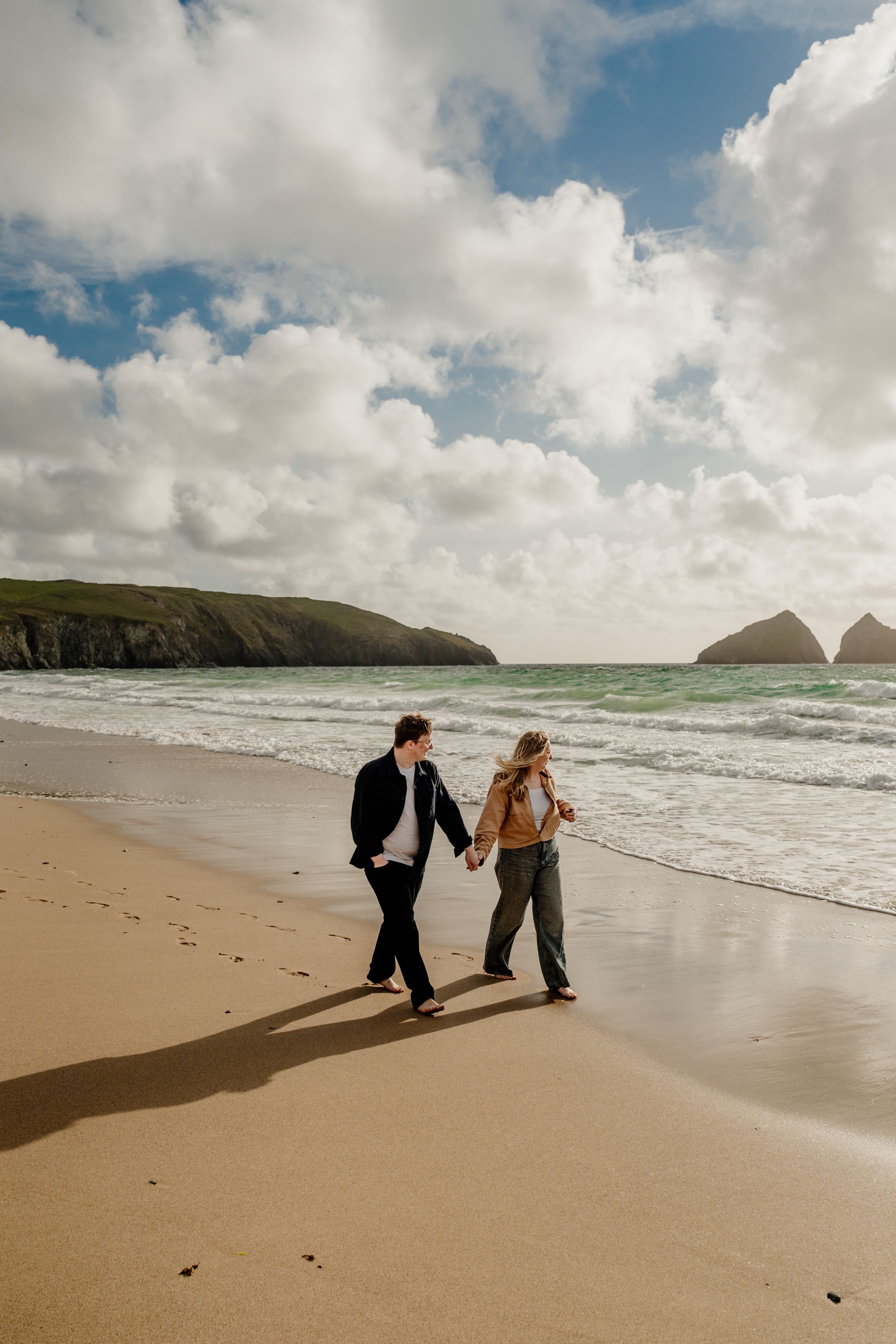 Couple holding hands walking on sandy beach with dramatic cloudy sky and rocky headlands in background.
