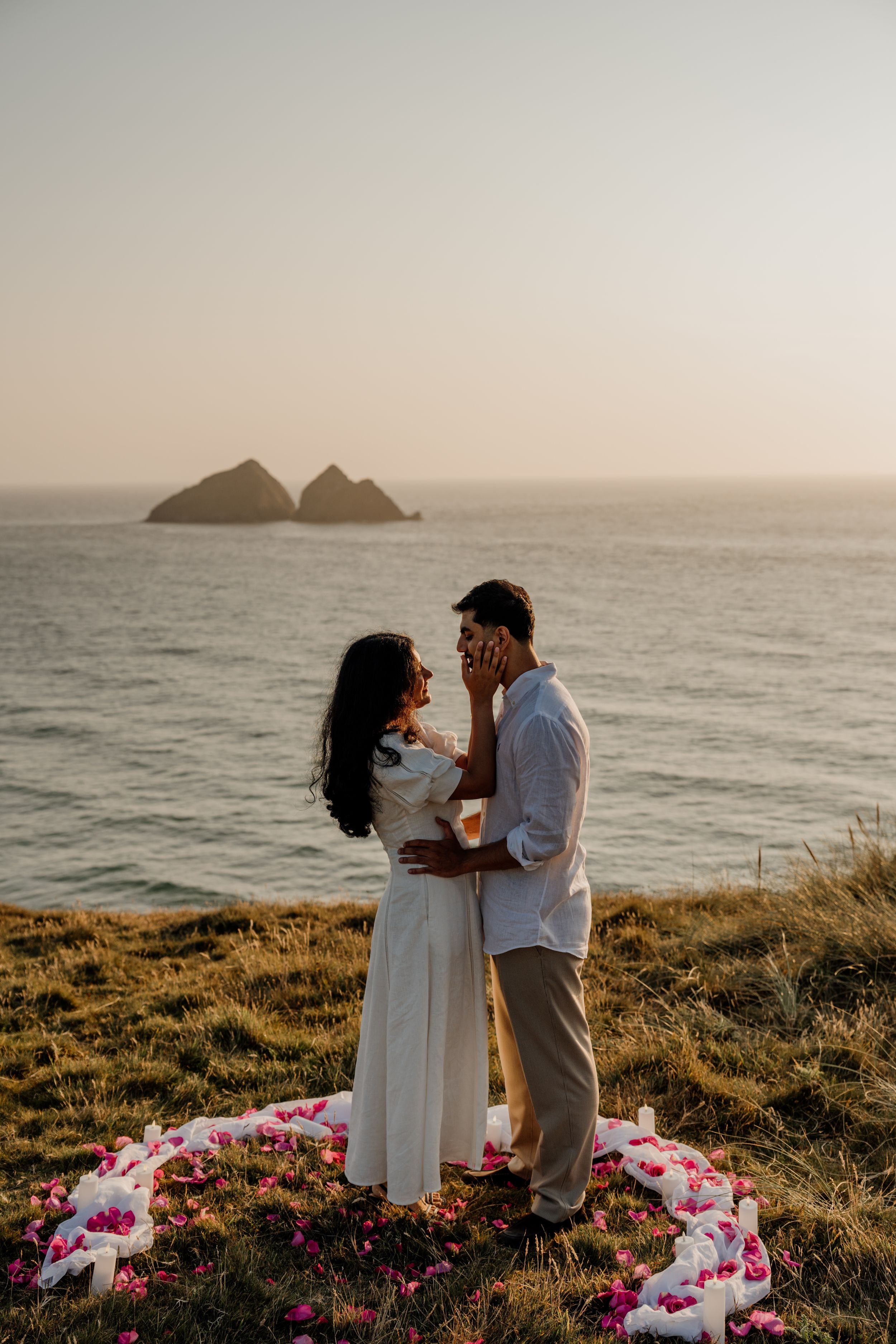 Couple sharing a romantic moment on a coastal cliff at sunset with flower petals and ocean rocks in the background.