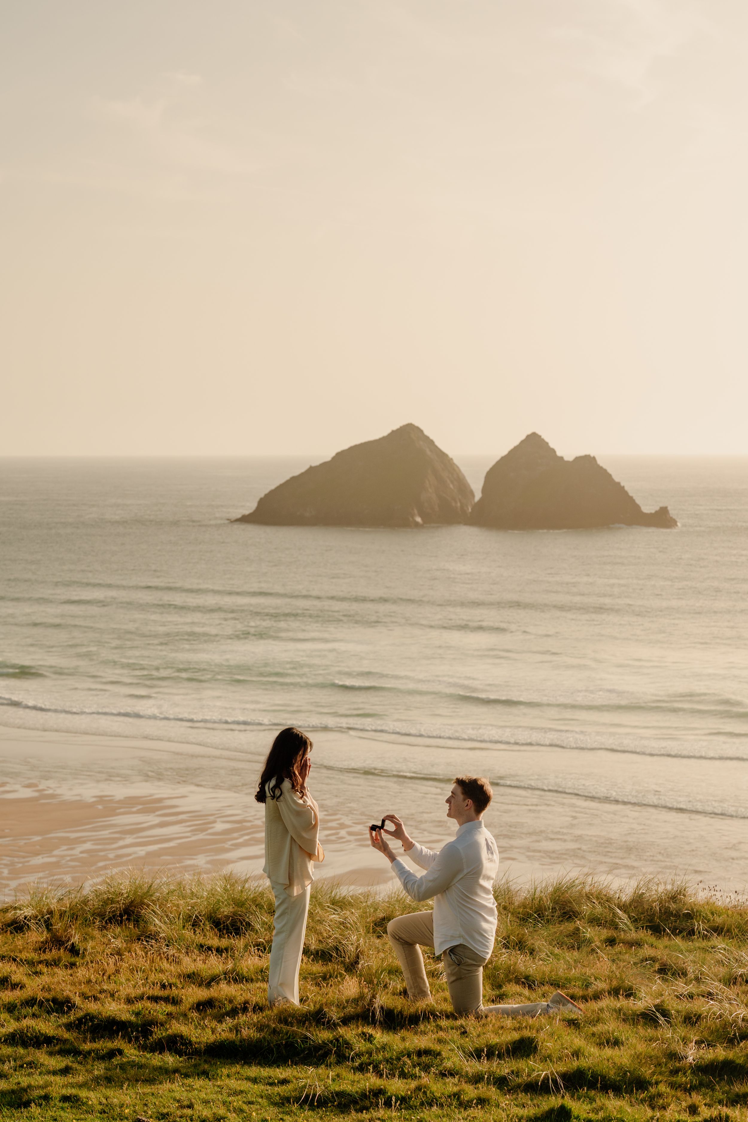 Man proposing to woman on grassy cliff overlooking sandy beach with twin rock formations at golden sunset.