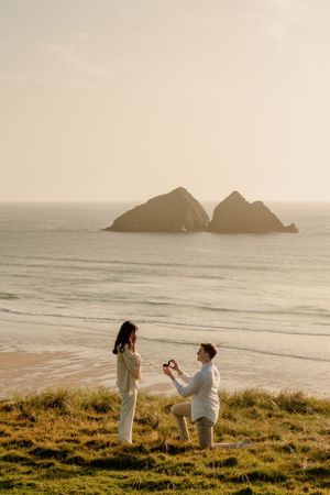 Man proposing to woman on grassy cliff overlooking sandy beach with twin rock formations at golden sunset.