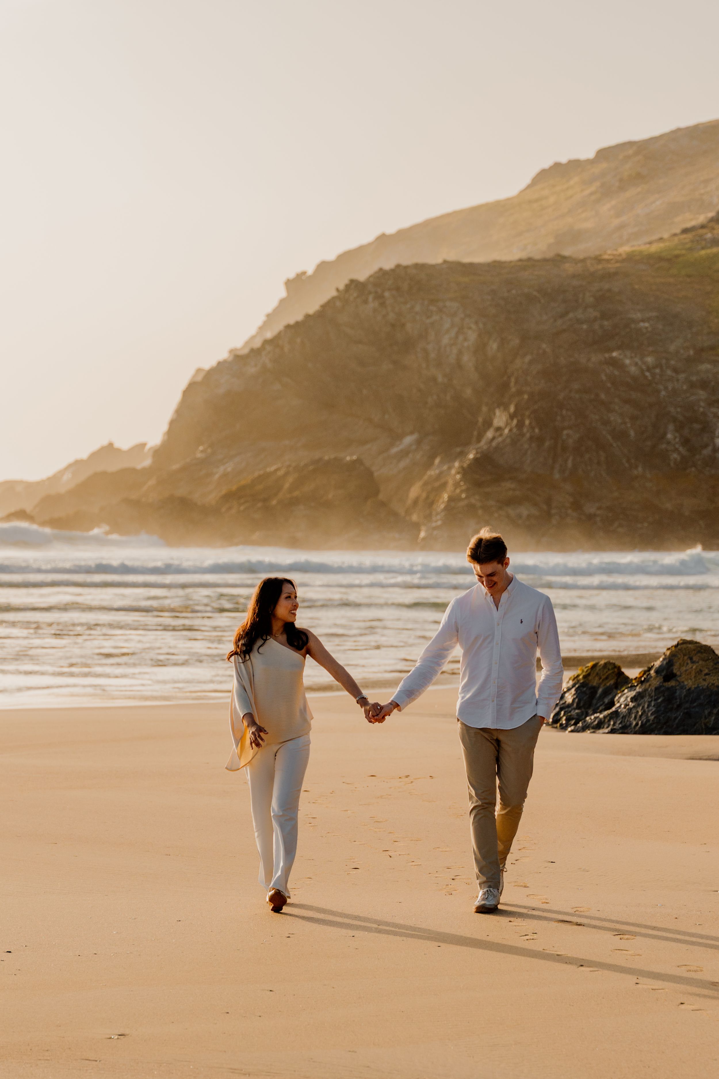 A couple holds hands walking on a sandy beach at golden hour with rocky cliffs and ocean waves in the background.