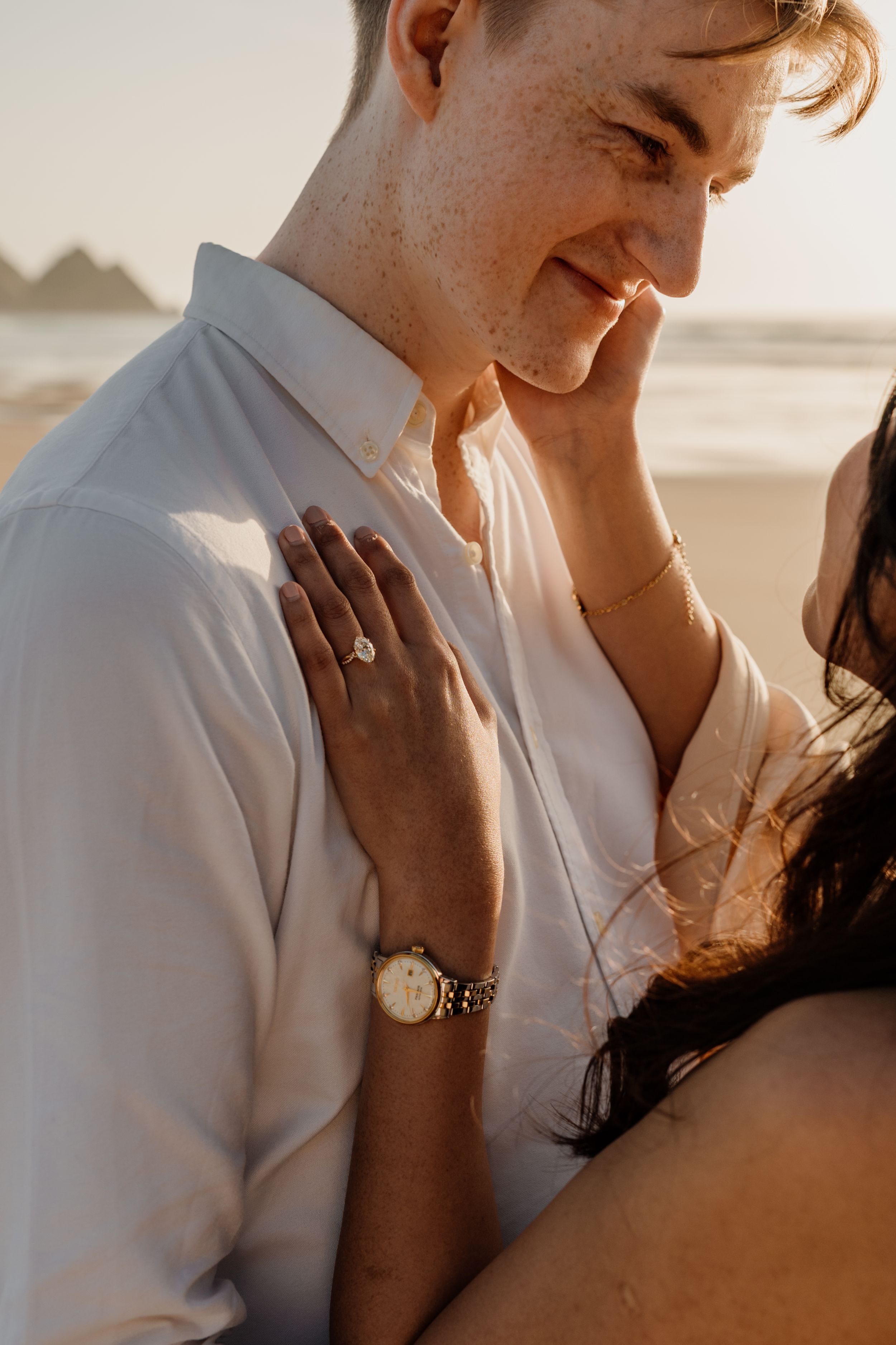 Smiling man with freckles in white shirt embraces woman on beach at golden hour, engagement ring visible.