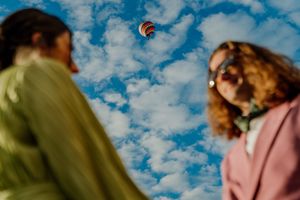 Two women look up at a colorful hot air balloon floating in a bright blue cloudy sky.