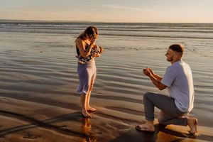 Man proposes to woman on a sandy beach at sunset, kneeling with a ring box as waves reflect golden light.