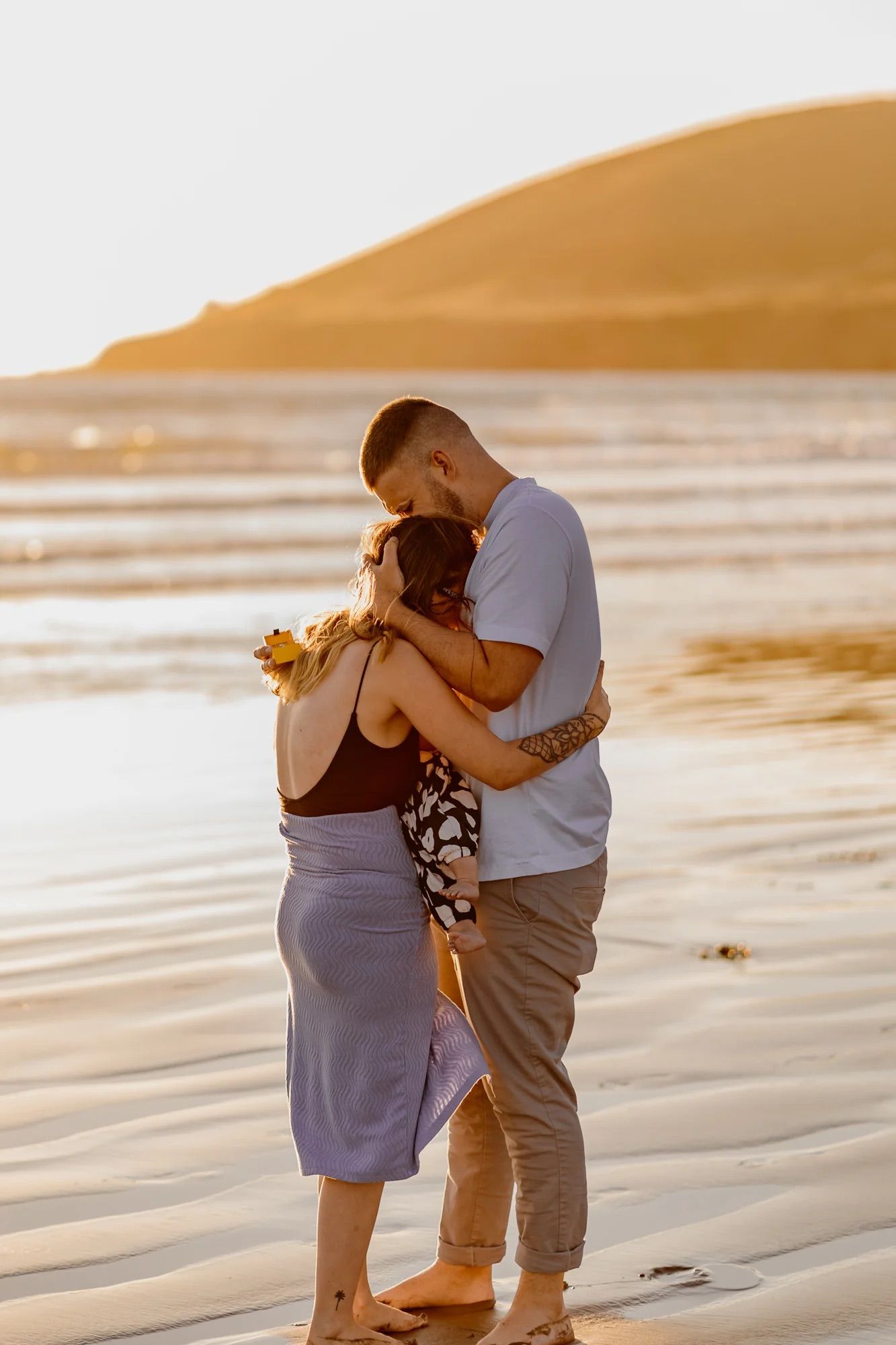 Couple embracing romantically on a sandy beach at golden sunset with ocean and hills in background.