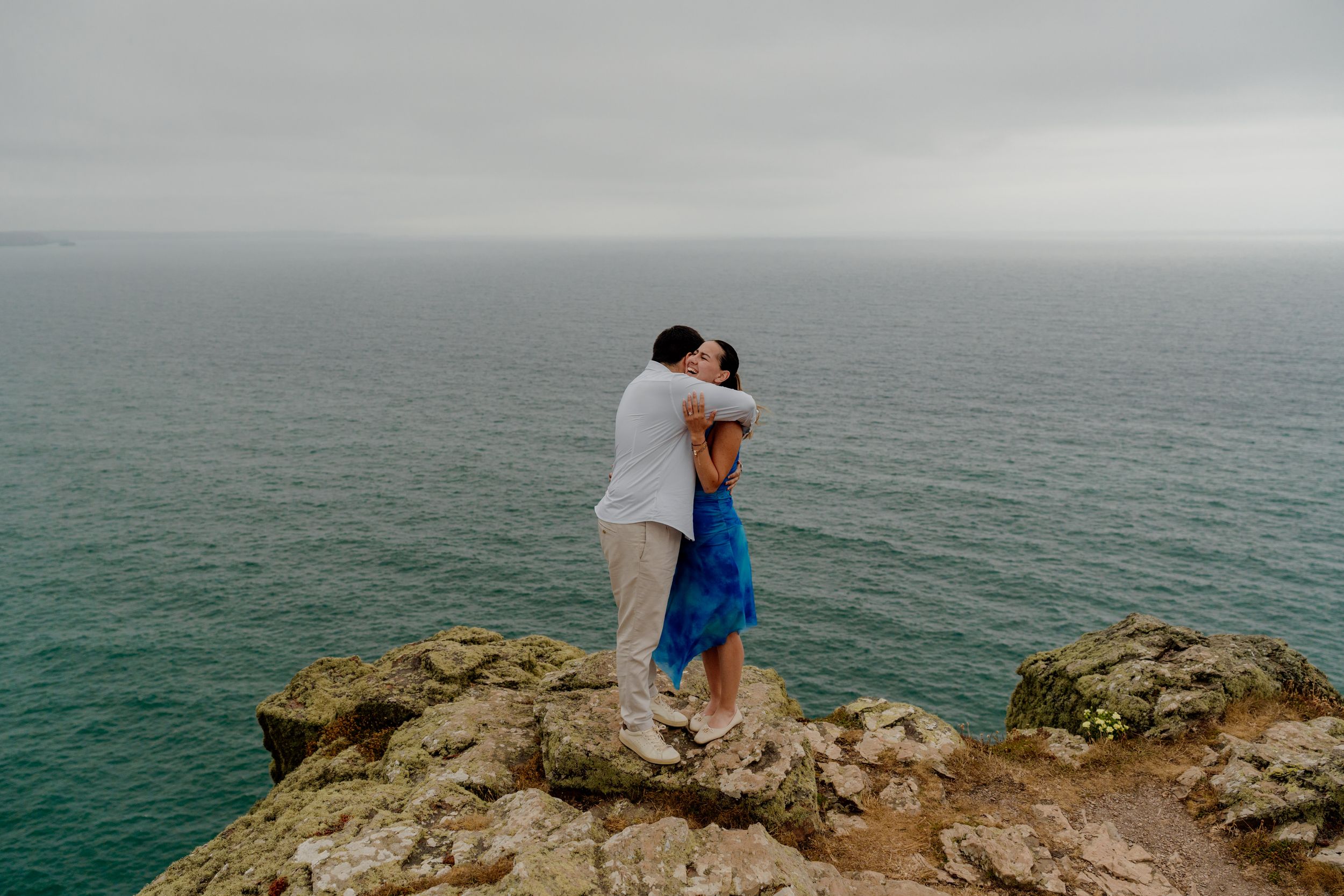 Couple embracing on rocky cliff overlooking turquoise ocean under overcast sky, woman in blue dress, romantic coastal scene.