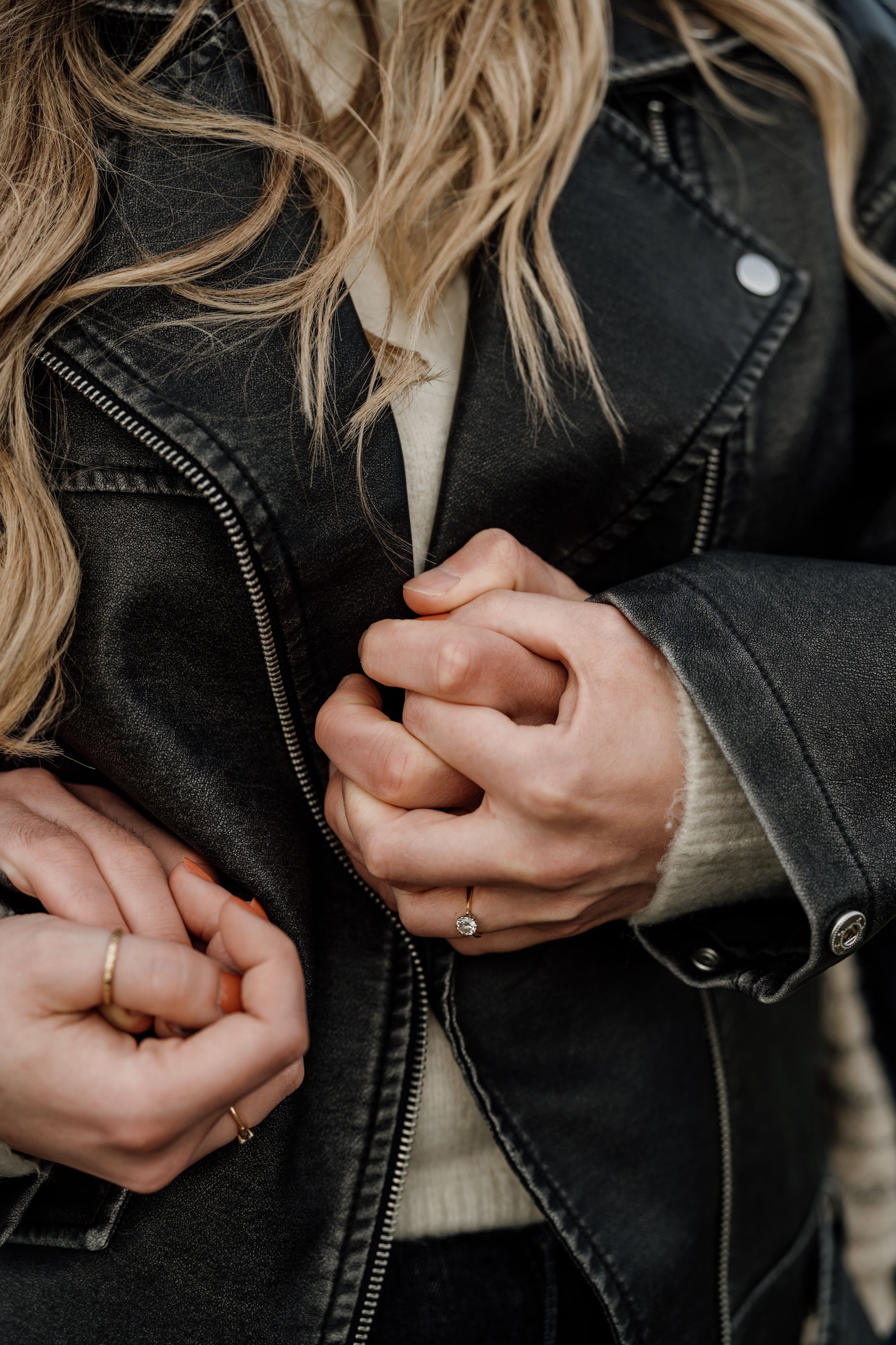Close-up of two people holding hands, wearing rings, with a black leather jacket and cream sweater visible.