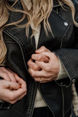 Close-up of two people holding hands, wearing rings, with a black leather jacket and cream sweater visible.