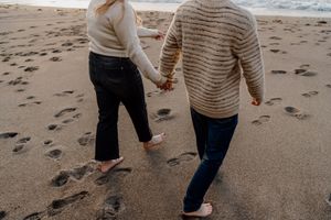 Couple holding hands walking barefoot on sandy beach leaving footprints in the wet sand.