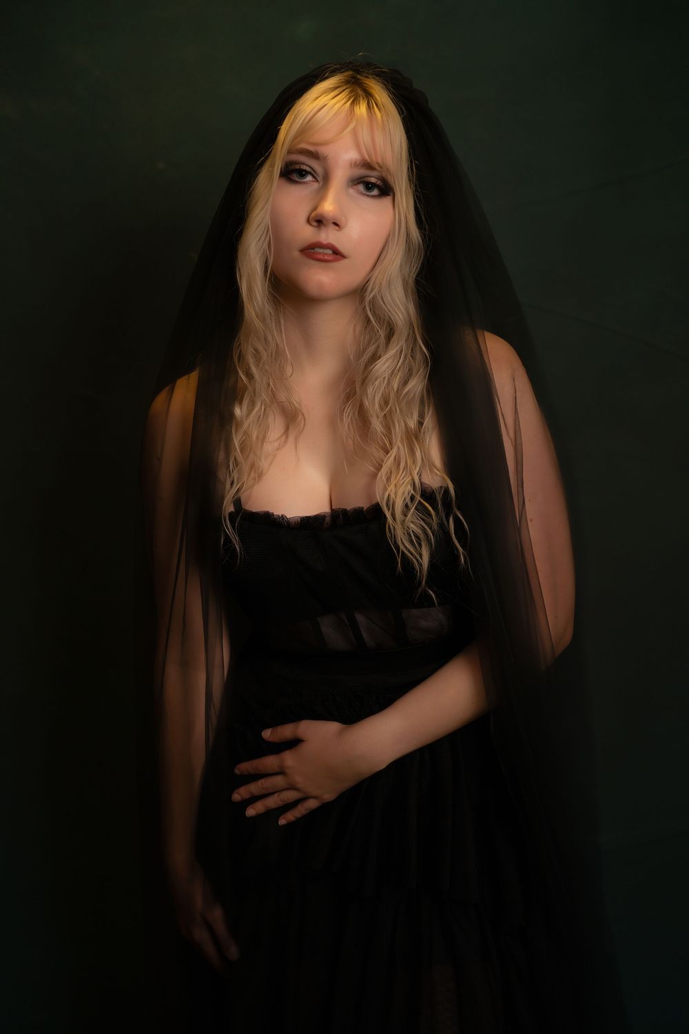 Young woman in black dress and veil with blonde hair poses against dark green background in gothic portrait.
