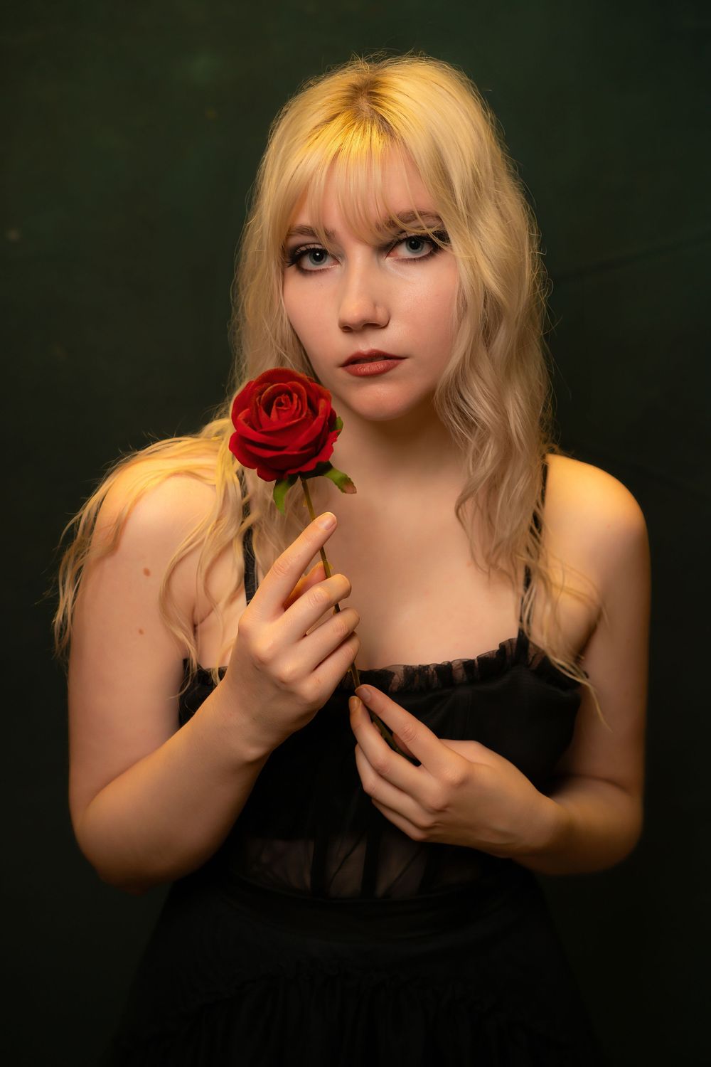 Young blonde woman in black dress holding a red rose, posing against dark green background in dramatic portrait.