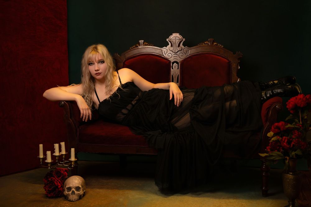 Blonde woman in black gown reclines on ornate red velvet sofa with candles, skull, and roses in gothic setting.