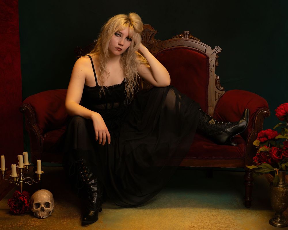 Young blonde woman in black gown poses on red velvet chaise with skull, candles, and roses in gothic-themed portrait.