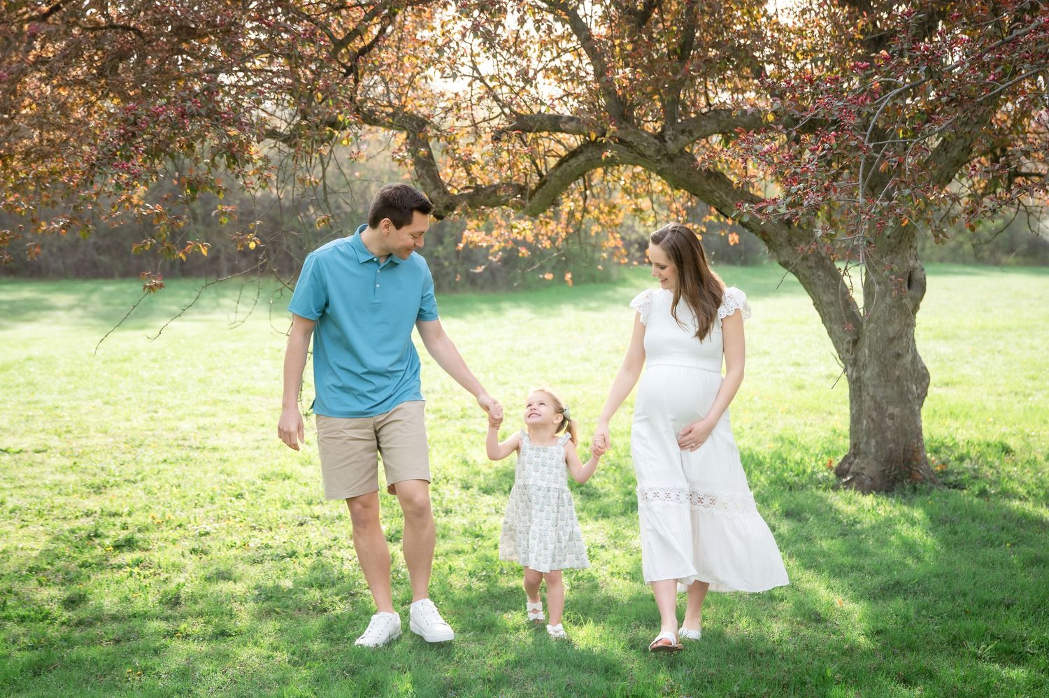 A pregnant woman in white dress holds hands with toddler girl and man in blue shirt walking in sunny park.