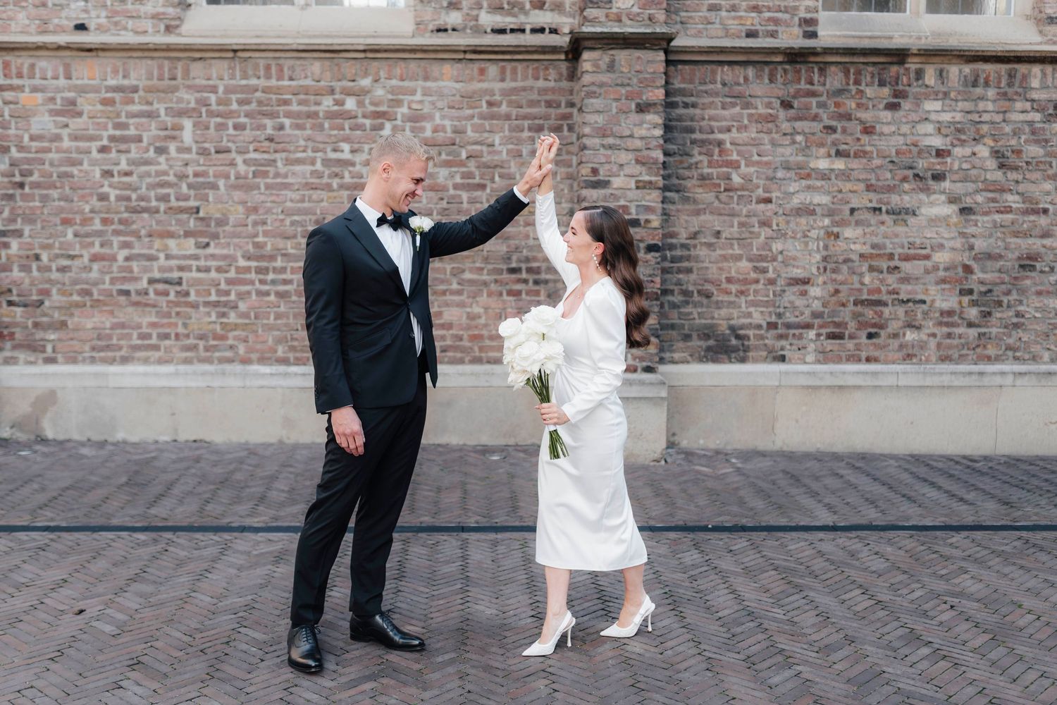 Bride and groom high-fiving in front of brick wall, wearing wedding attire, bride holding white bouquet.