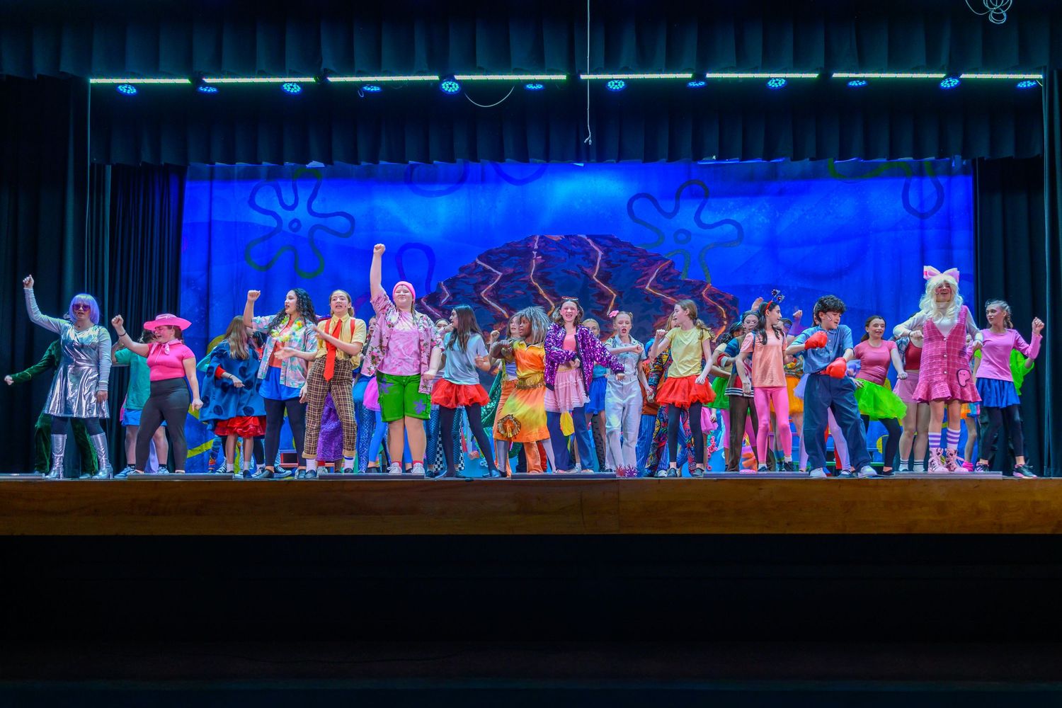 Colorful school stage performance with children in bright costumes performing a musical in front of a blue ocean backdrop.