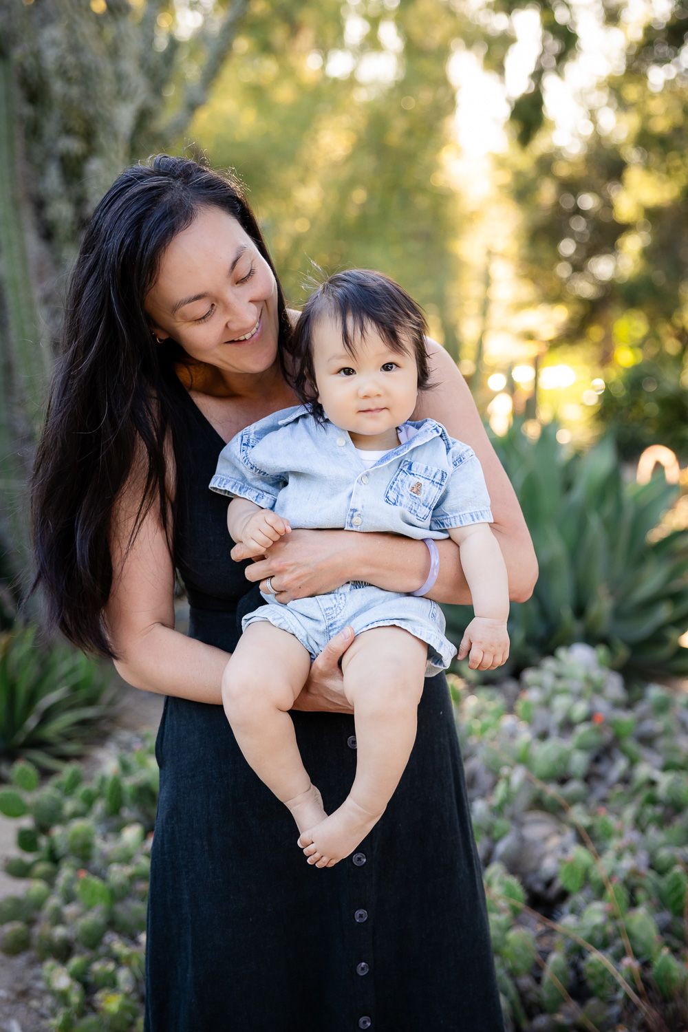 A smiling mother in a black dress holds her baby boy in a denim outfit outdoors in a lush garden in Palo Alto, CA
