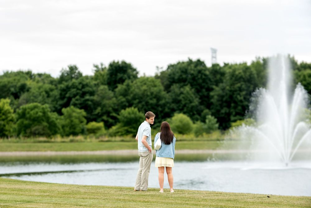Couple holding hands facing a serene pond with a fountain, surrounded by lush green trees in a park.