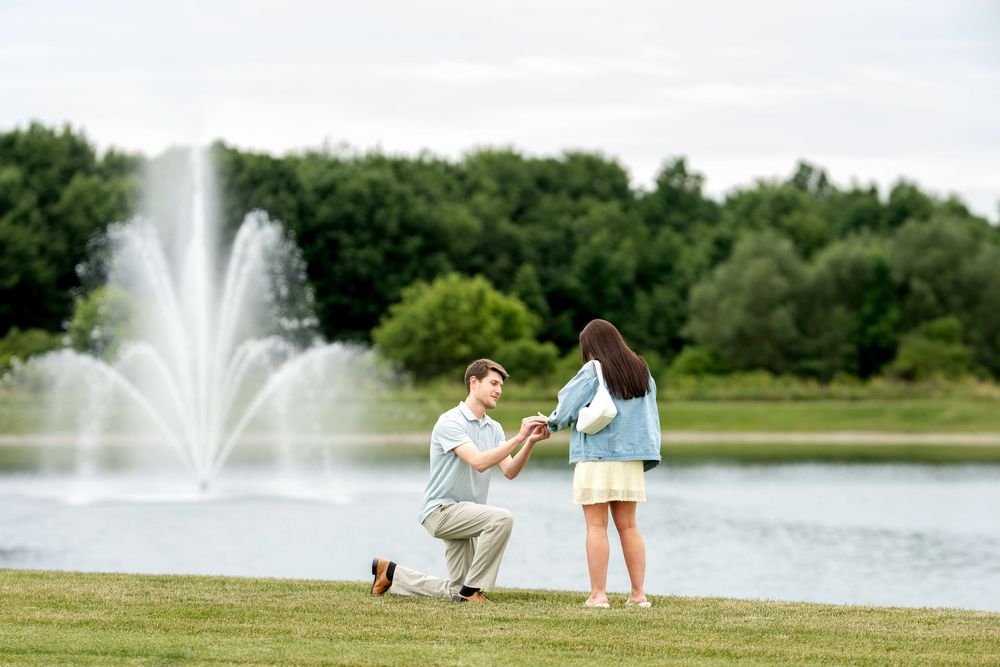Romantic marriage proposal by a lake fountain, man on one knee presenting ring to his girlfriend outdoors.