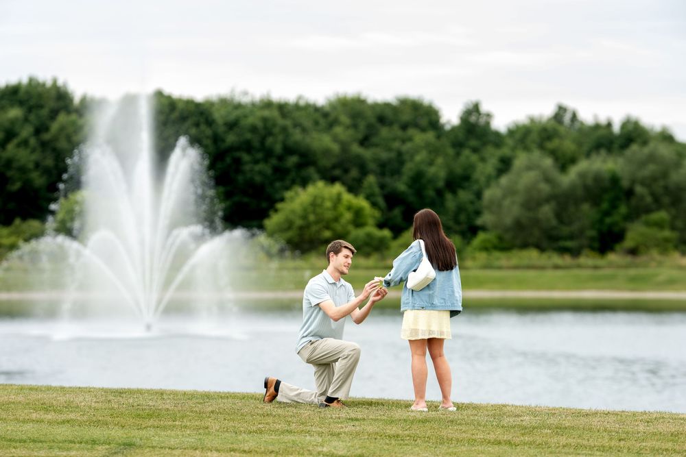 Man on one knee proposing to woman with ring near a fountain and lake in a green park.
