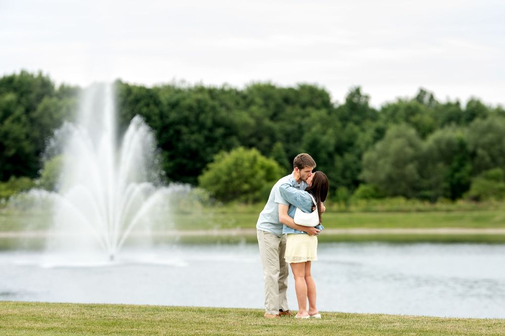 Couple sharing a romantic kiss near a beautiful fountain by a serene lake in a green park.