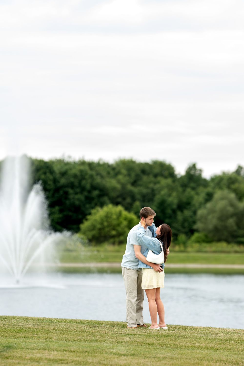 Romantic couple embracing near a serene park fountain with lush green trees in the background on a cloudy day.