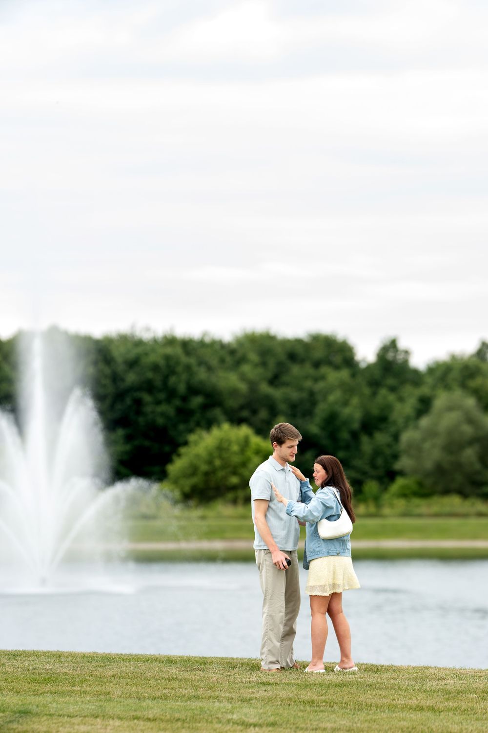 Couple standing together by a serene park pond with a fountain, surrounded by lush green trees on a cloudy day.