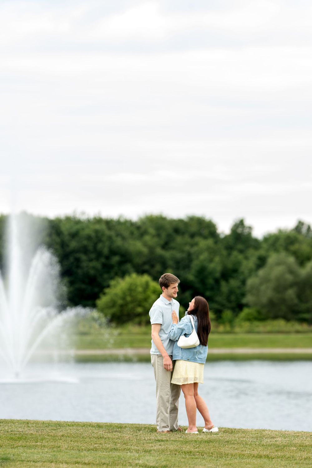 Young couple holding hands and facing each other by a decorative park fountain surrounded by green trees.