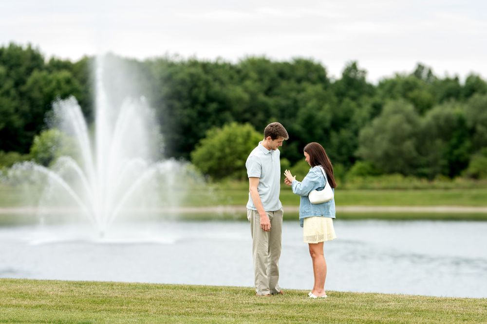 Couple standing together examining engagement ring near a decorative fountain pond in a green park.