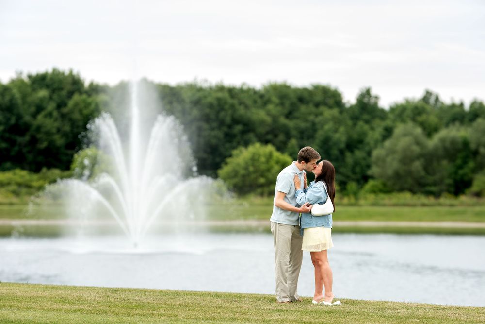 Newly engaged couple sharing a kiss by a beautiful pond with a large water fountain and green trees.