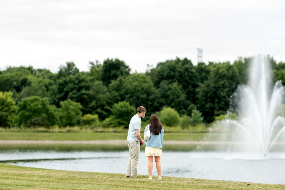 Couple holding hands by a serene pond with a fountain and lush green trees in a park.