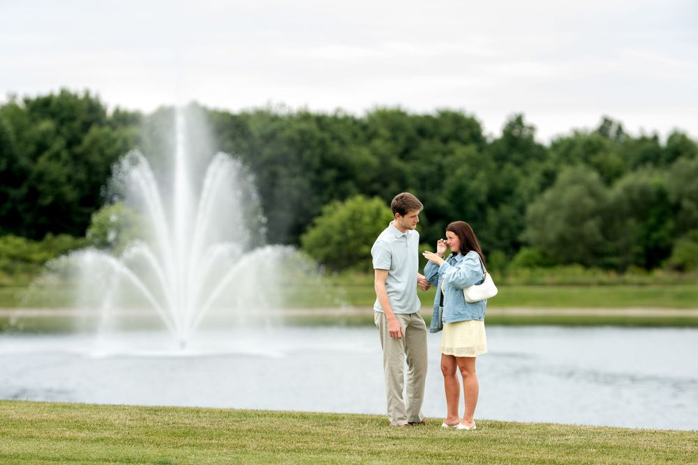 Happy engaged couple admiring engagement ring together beside a scenic lake fountain in a park.