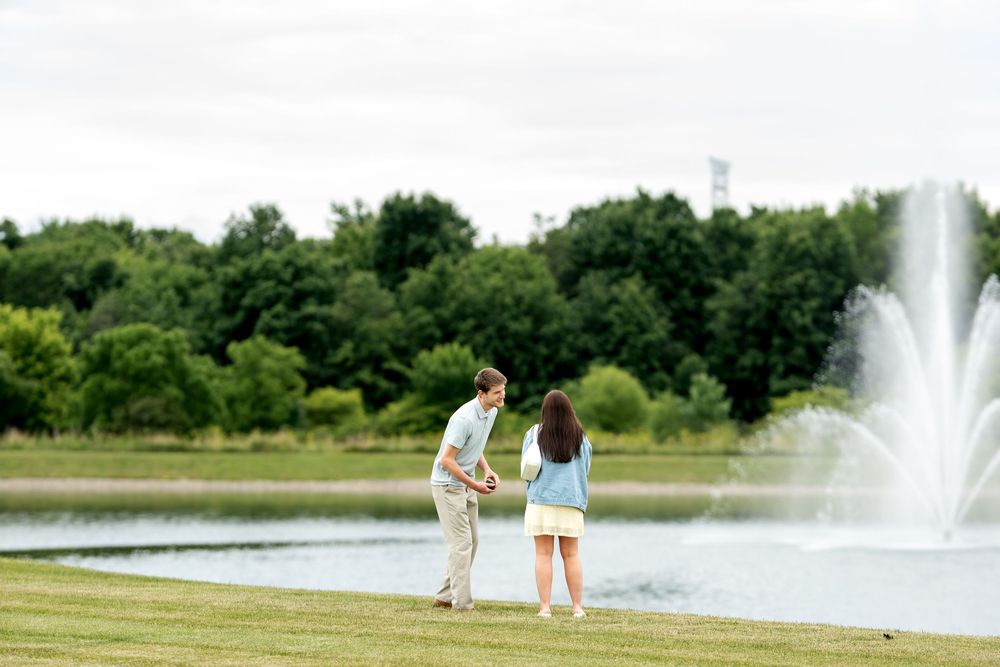 Couple standing by a serene park lake with a fountain, surrounded by lush green trees on a cloudy day.