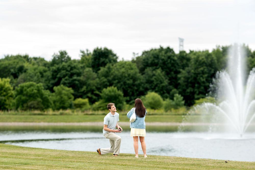 Man kneeling to propose to woman by a scenic pond with fountain and lush green trees in background.