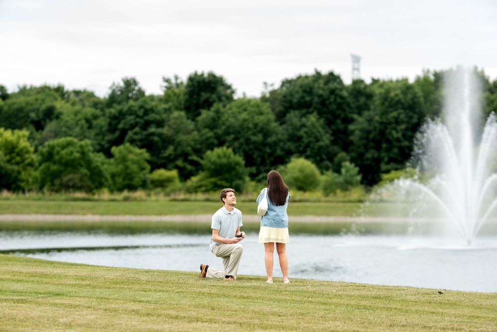 Man proposing on one knee to woman by a scenic pond with fountain in a lush green park.