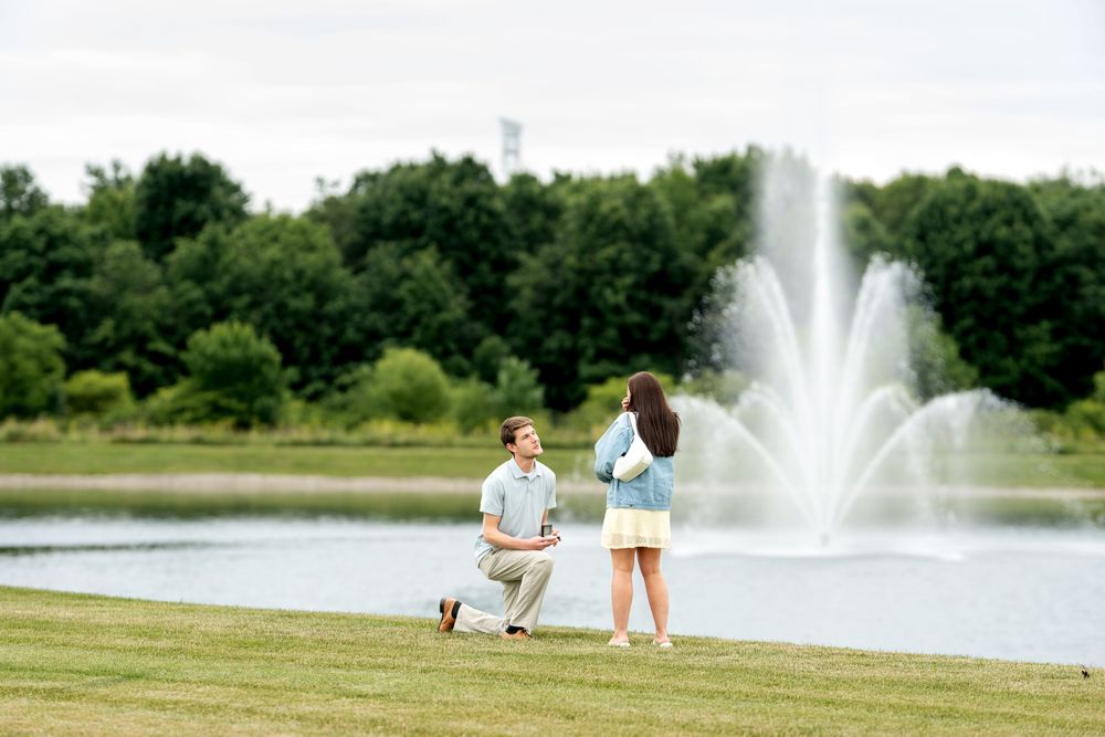 Romantic marriage proposal on one knee near a fountain pond in a beautiful outdoor park setting.