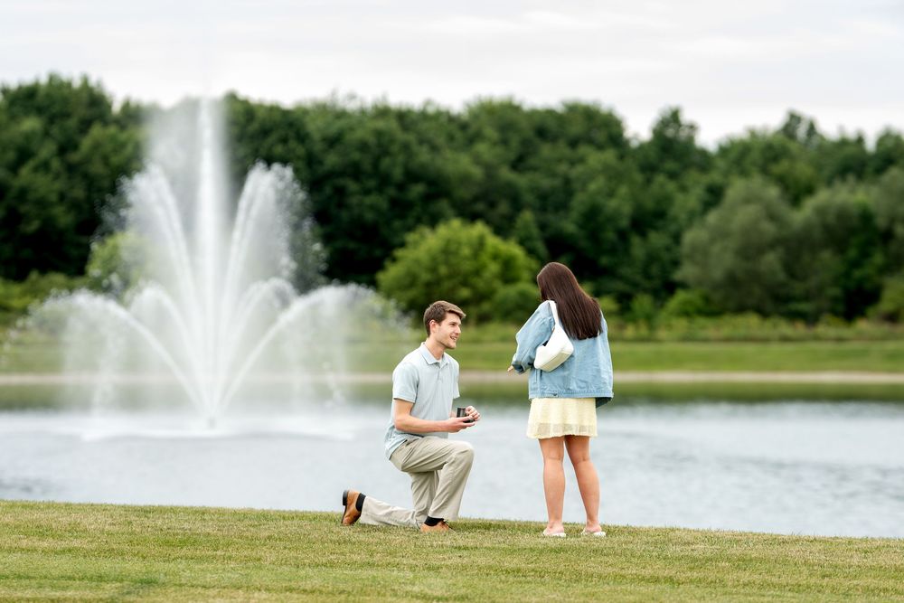 Man proposing on one knee to woman by a pond with a fountain in a green park setting.