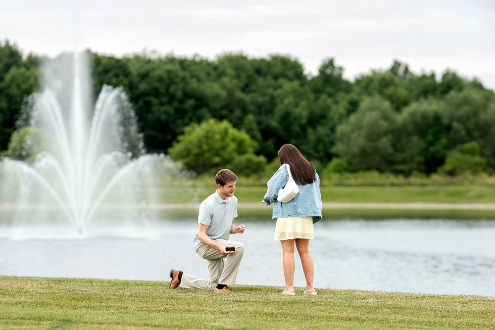 Man proposing on one knee to woman by a scenic pond with fountains in a lush green park.