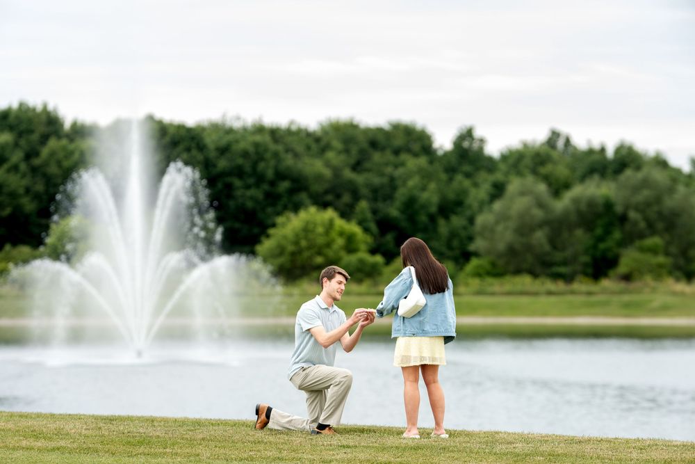 Man proposes to woman on one knee by a scenic pond with a fountain and lush green trees in background.